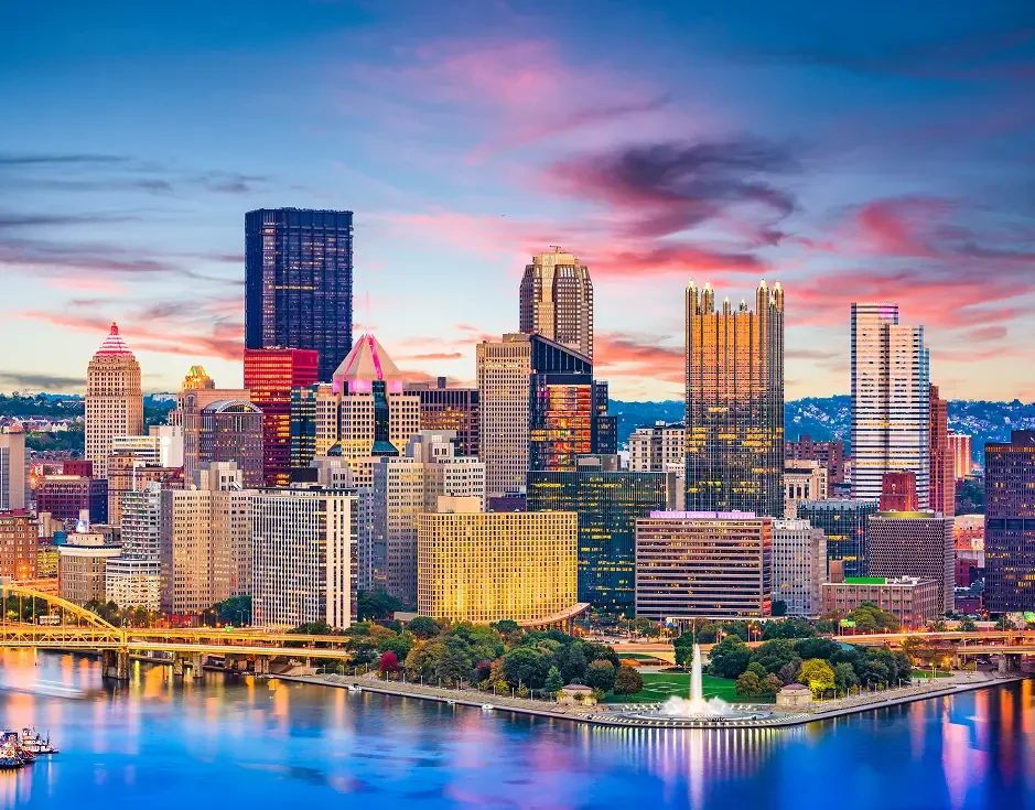 Pittsburgh skyline at dusk, showing prominent skyscrapers and a river in the foreground.
