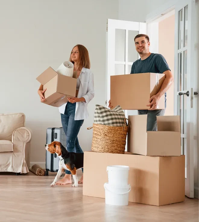 A couple moving into a new home, carrying cardboard boxes while their dog stands nearby in a bright living room filled with packed belongings.