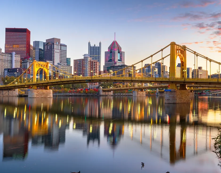Golden bridge spanning a calm river with city skyline and colorful sunset sky reflecting on the water.