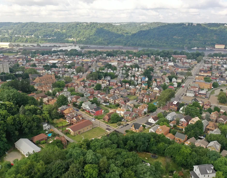 Aerial view of a small town with dense houses, green trees, a river, and forested hills in the background under a cloudy sky.