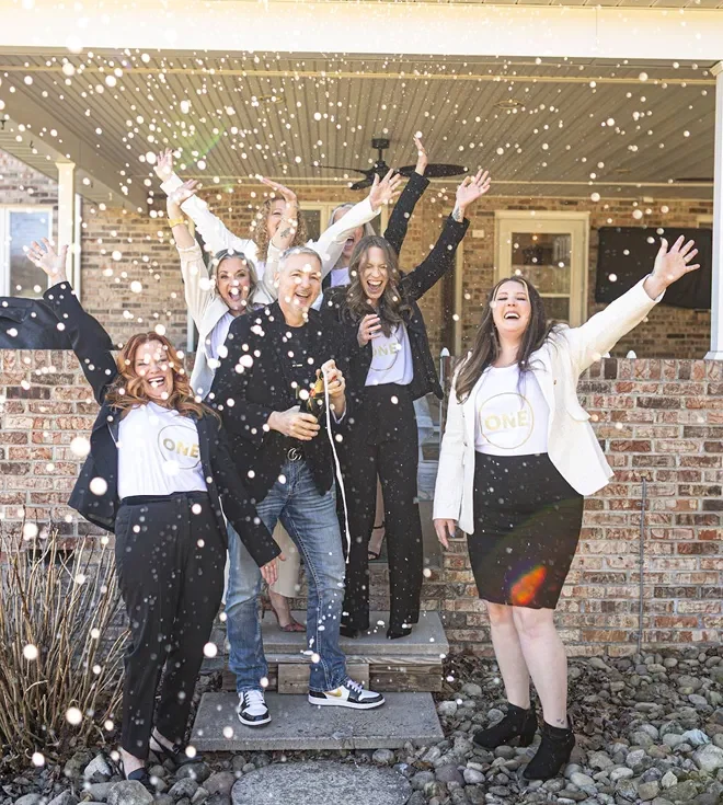 A group of seven professionals from The Moio-Iseman Group in business casual attire celebrating with confetti outside a brick house
